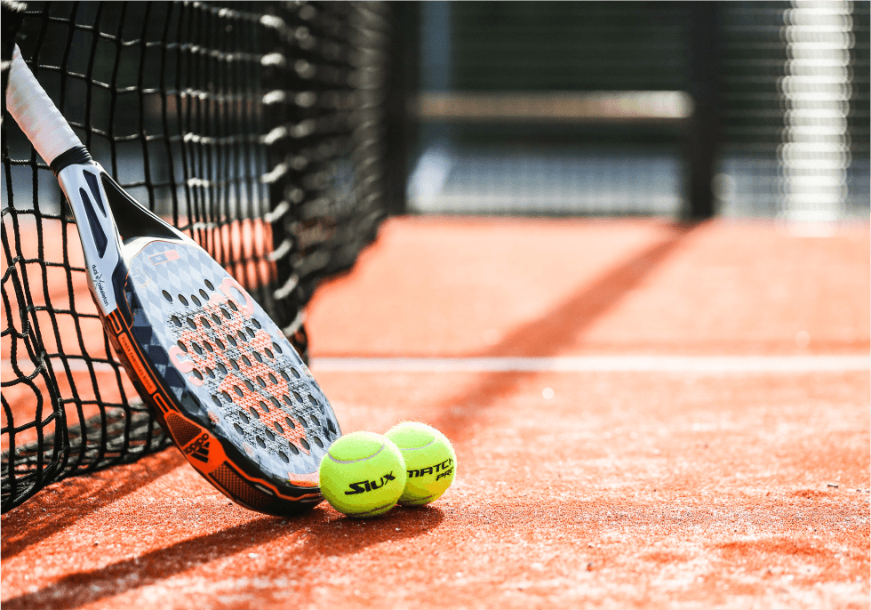 padel paddle leaning against the net surrounded by balls on an orange court